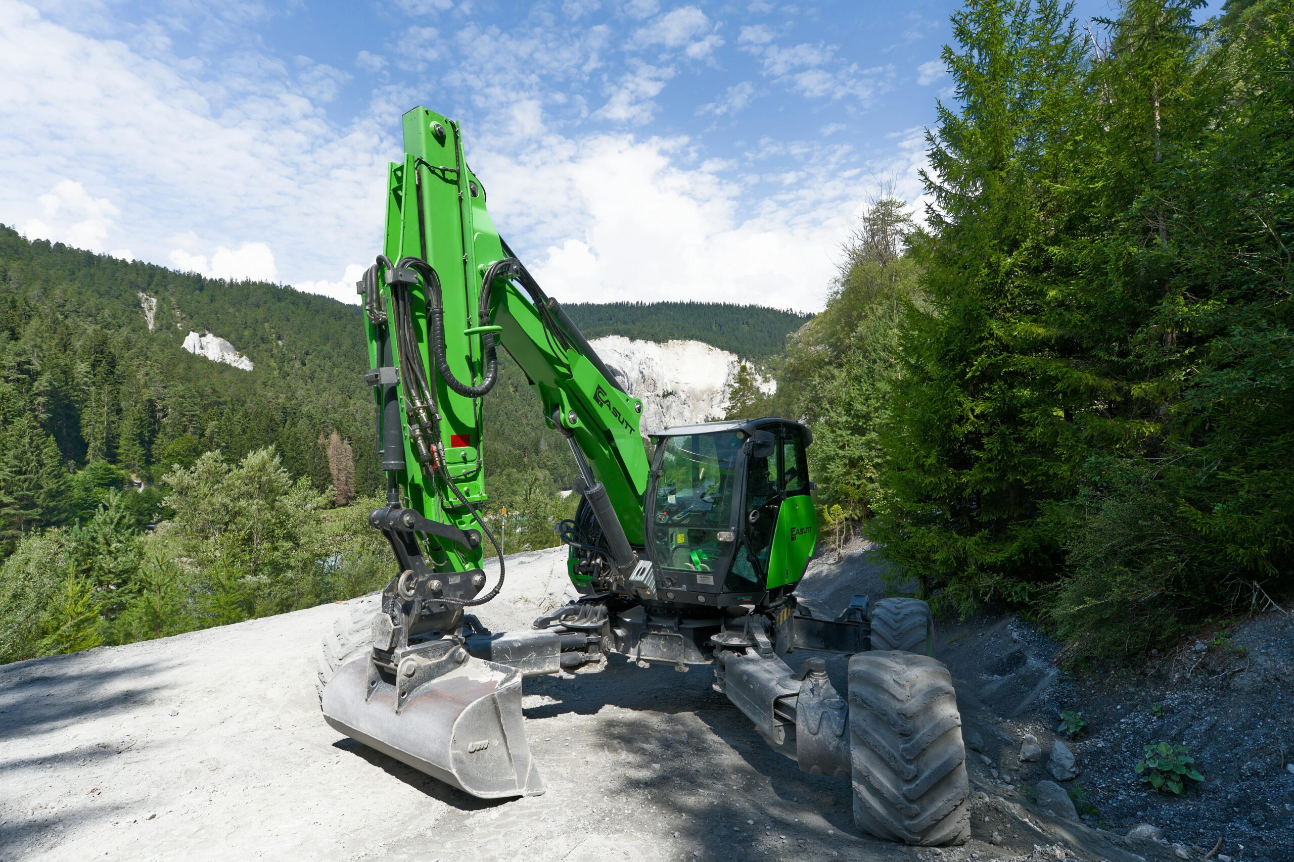 Home A green excavator operating in a scenic Swiss alpine forest with clear skies.